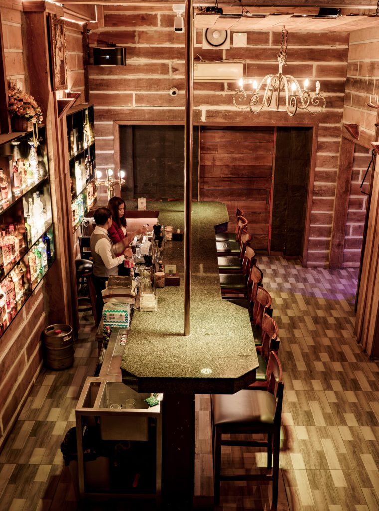 Narrow bar interior with wooden walls, high tables, and shelves stocked with bottles along the side.