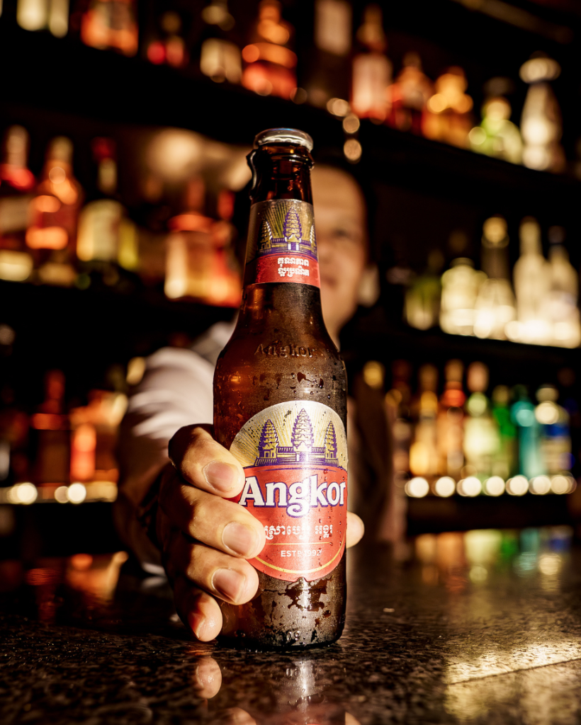 San Miguel beer bottle on a bar counter with warm lighting and a blurred bar shelf in the background.